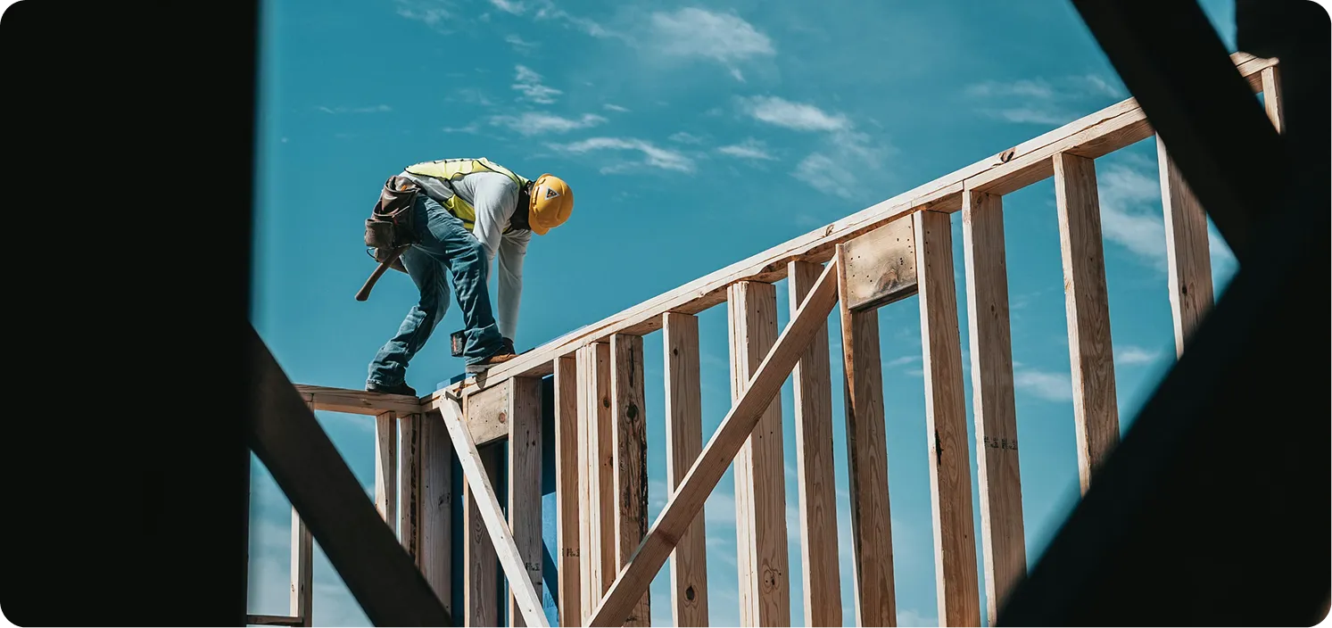 Construction worker installing wooden framing on a building structure under a clear blue sky.